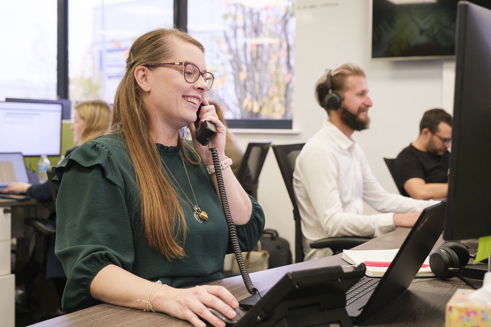 A lady working as a Managed IT supplier talking on a phone in front of a desktop PC in an office with people in the background.