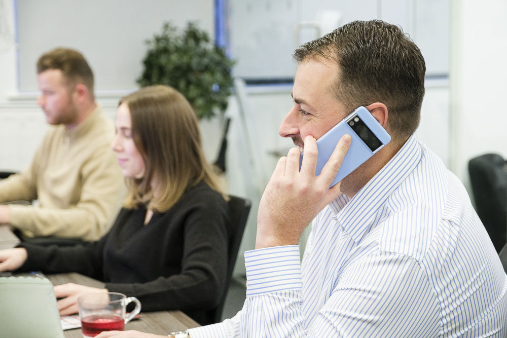 Man in office talking on his business mobile phone with other employees in the background.